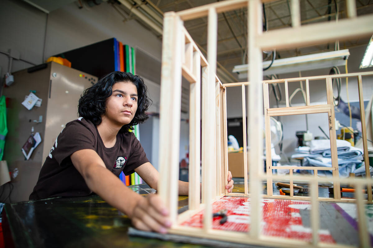 A young person with dark, wavy hair assembles a wooden architectural model in a workshop, focusing intently on aligning the structure. Colorful materials and equipment are visible in the background.
