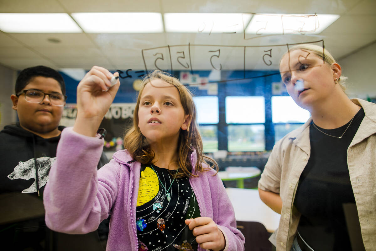 Three students stand in a classroom, with one girl writing on a transparent board using a marker, while the others watch her work attentively. The classroom is bright and filled with natural light.