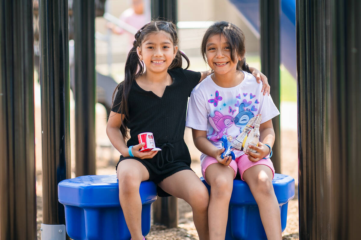 Two young girls sit side by side on blue playground seats, smiling at the camera. One wears a black outfit and holds a soda can; the other wears a white shirt with cartoon characters and holds snacks.