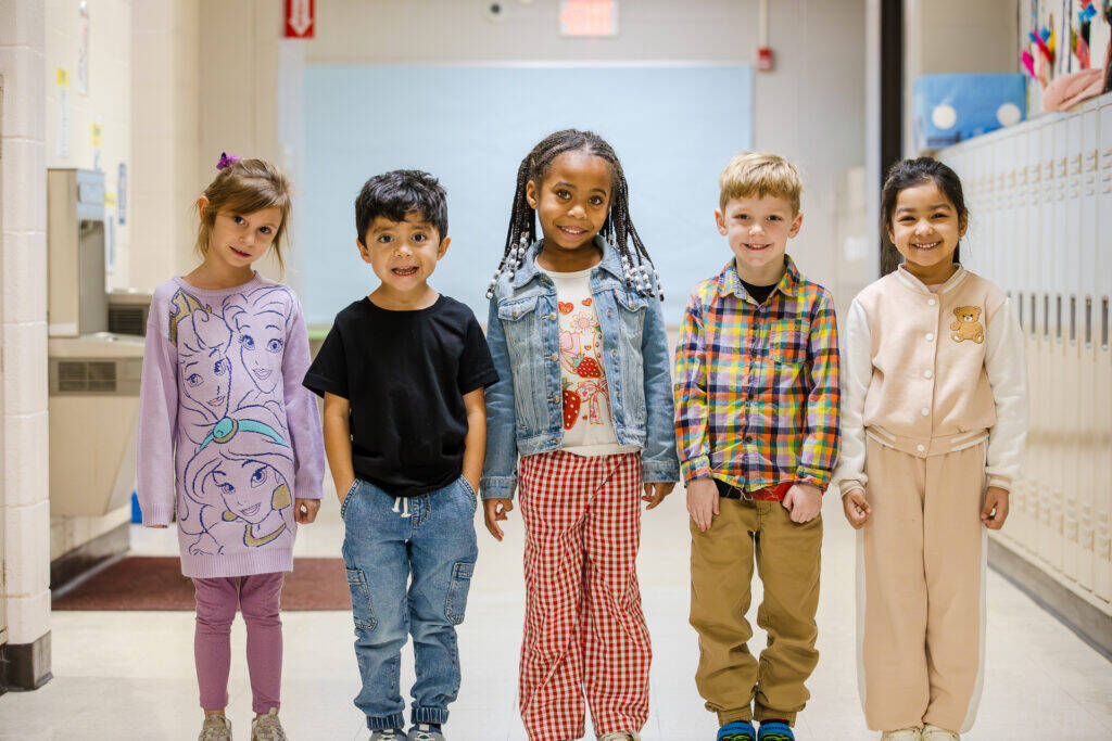 Five young children stand in a school hallway, smiling and holding hands. They wear colorful, casual clothes and are positioned in front of lockers and a light blue bulletin board.