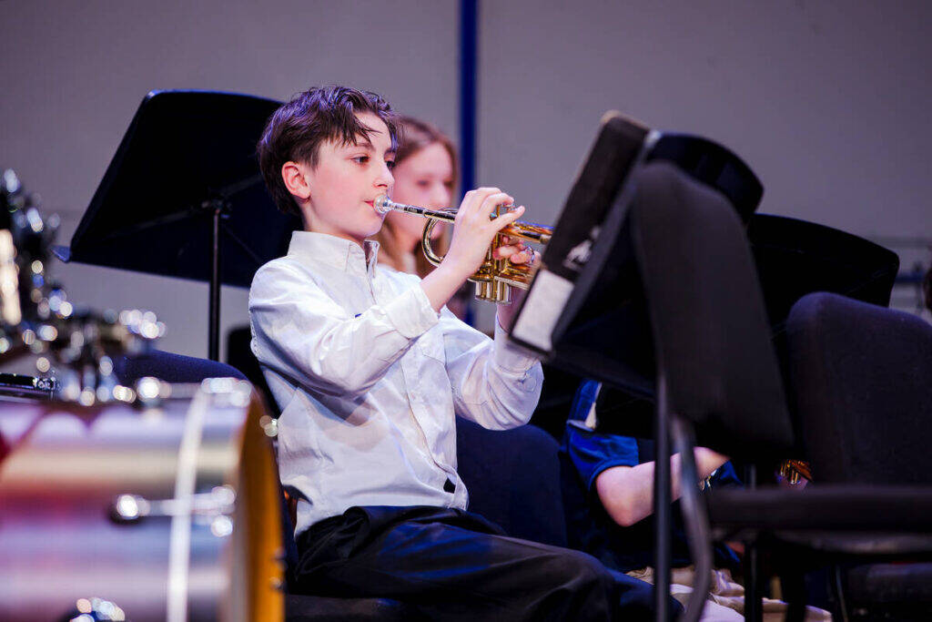 A young boy in a white shirt plays the trumpet while seated on stage during a music performance, with other musicians and music stands visible in the background.