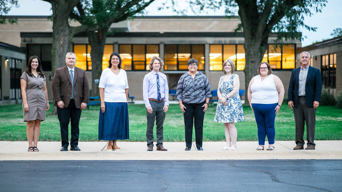 Eight adults stand spaced apart in a row on a sidewalk in front of a building with large windows, trees, and grass. The group includes men and women dressed in business and casual attire.