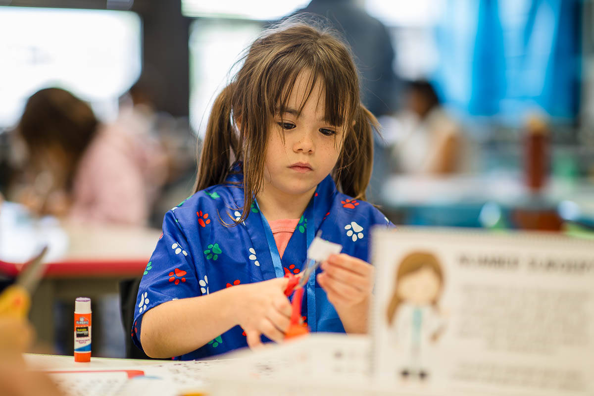 A young girl in a blue floral shirt sits at a table, using scissors and paper for a craft activity, with a glue stick nearby.